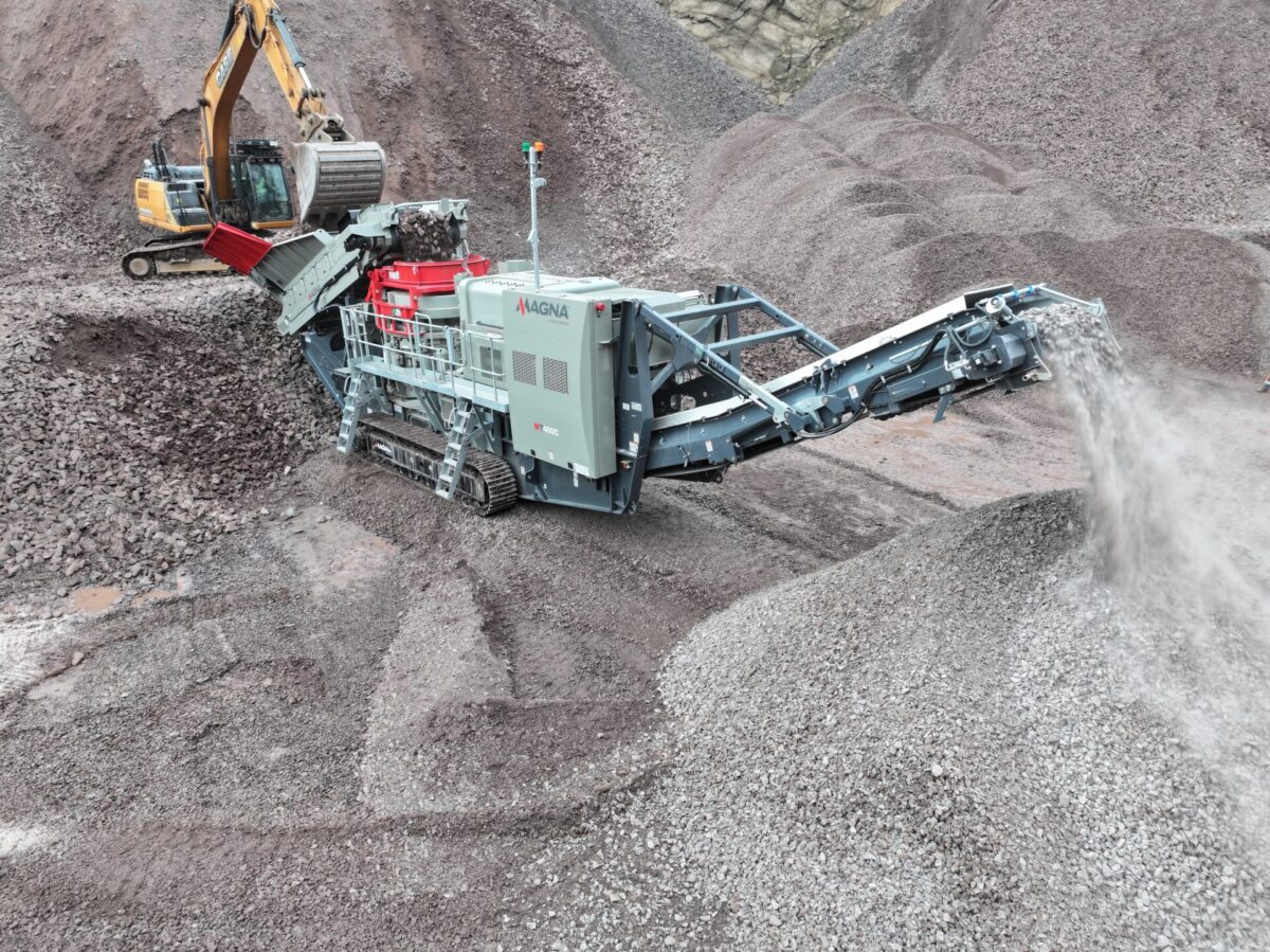 MAGNA heavy-duty cone crusher in operation with an excavator feeding raw rock, producing clean aggregate stockpiles in a quarry setting.