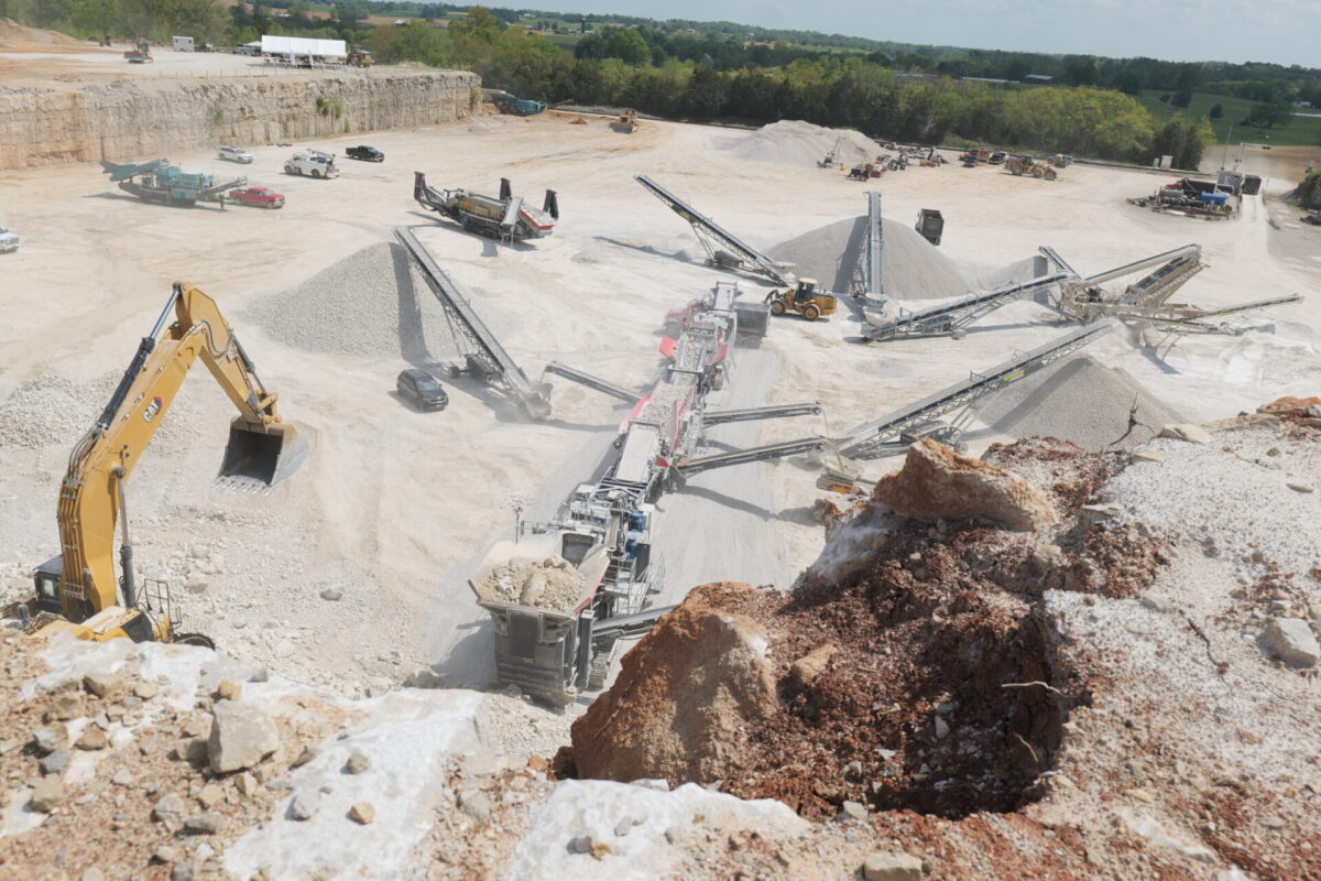 Aerial view of a large quarry operation featuring multiple MAGNA crushers, screeners, and conveyors processing rock, with an excavator loading material into a mobile crusher and stockpiles of gravel surrounding the site.
